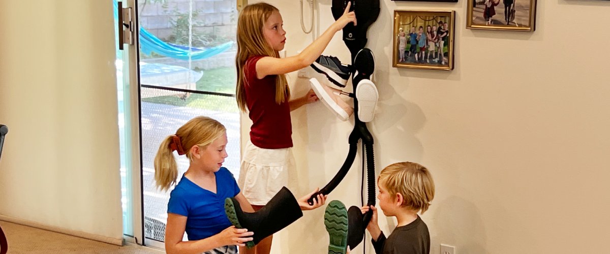 Bērni priekšnamā izmanto pie sienas piestiprinātu apavu žāvētāju, novietojot slapjus zābakus un apavus žāvēšanai pēc atgriešanās no āra