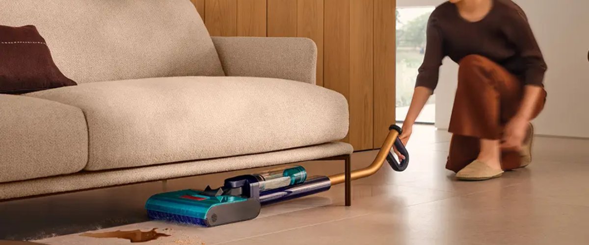 Sieviete ar Dyson WR03 cieto grīdu tīrītāju tīra zem dīvāna izlijušu šķidrumu un netīrumus