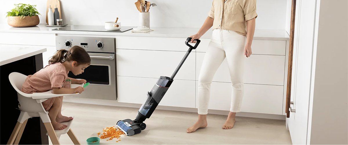Sieviete tīra virtuves grīdu ar bezvadu mazgājošo putekļu sūcēju, kamēr mazs bērns sēž barošanas krēslā un izber ēdienu uz grīdas.