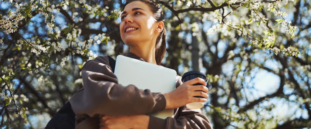 Jauna sieviete tur rokās MacBook un kafijas krūzi, stāvot pavasarīgā, ziedošā parkā