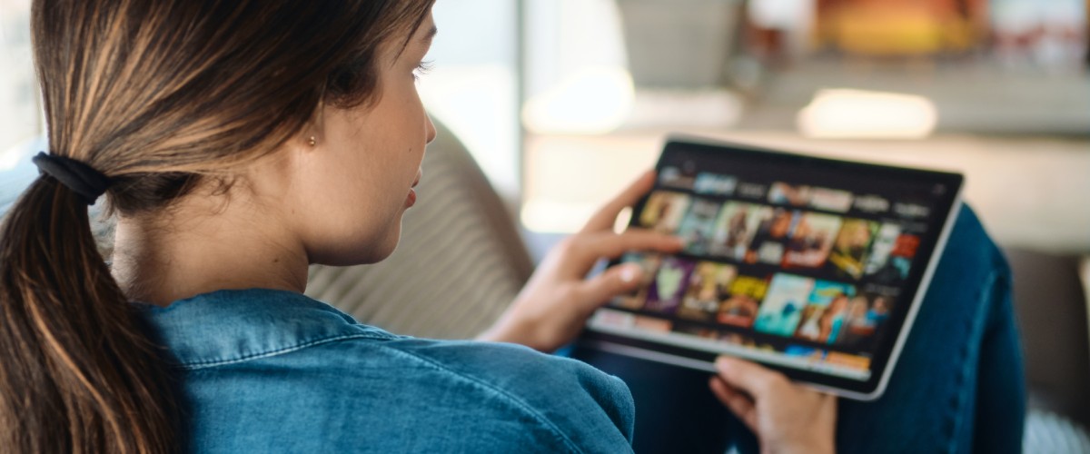 Sieviete mājās uz dīvāna planšetdatorā pārlūko straumēšanas pakalpojuma filmas un seriālus