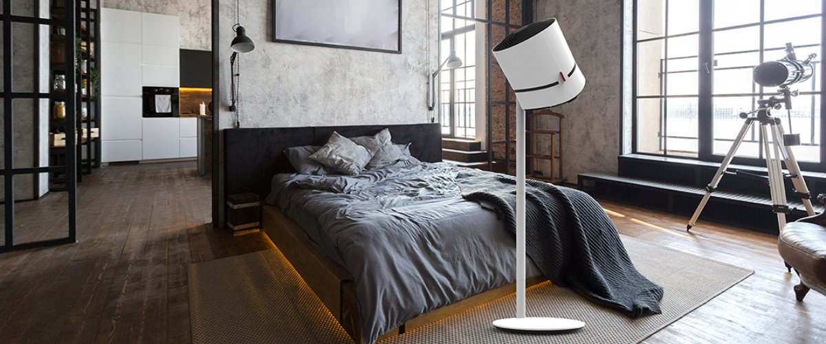Balts grīdas ventilators pie gultas plašā lofta stila guļamistabā ar lieliem logiem