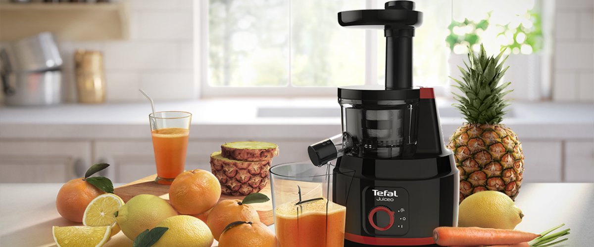 Tefal Juiceo lēnās darbības sulu spiede uz virtuves darba virsmas, blakus svaiga sula, citrusaugļi, ananass un burkāni